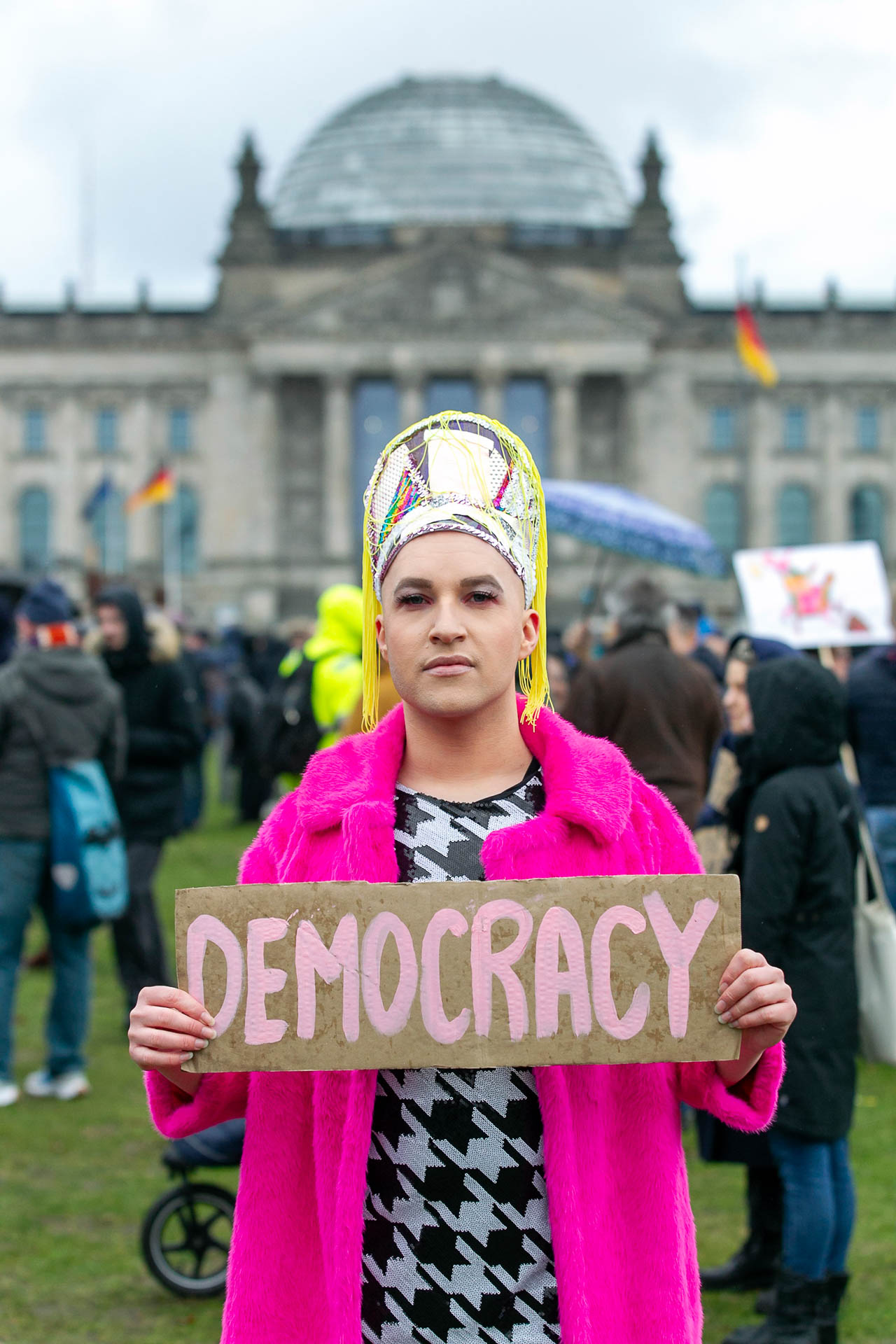 Mitglied einer Demonstration in auffälliger Kleidung – einem pinken Kunstpelzmantel und einer kunstvollen Kopfbedeckung – die ein Schild mit der Aufschrift "DEMOCRACY" in pinker Schrift hält. Im Hintergrund ist das Reichstagsgebäude in Berlin zu erkennen