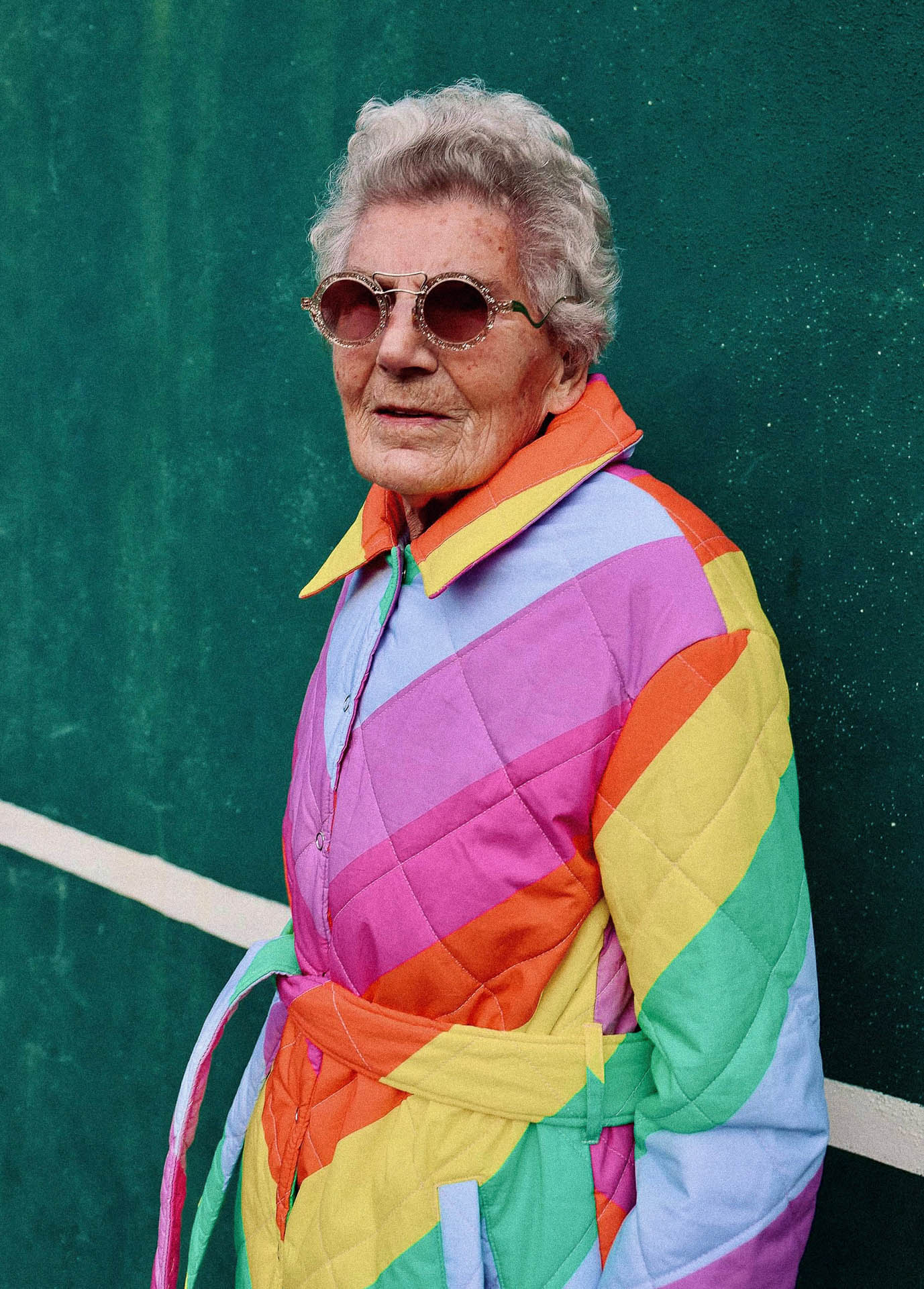 Ältere Dame mit Sonnenbrille und bunt gestreiftem Mantel vor einer dunkelgrünen Wand.