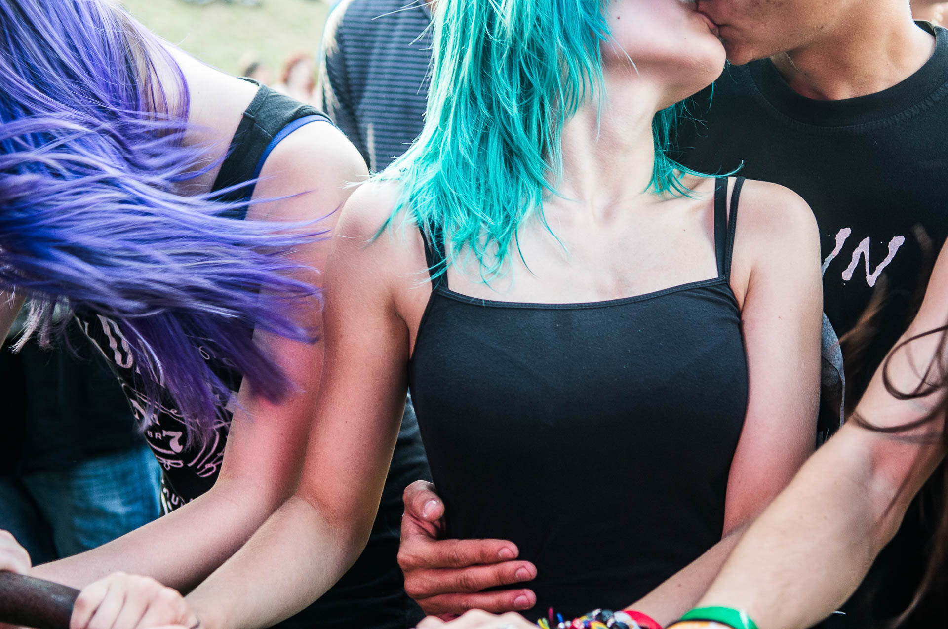 Das Bild zeigt eine lebendige und emotionale Szene, vermutlich auf einem Konzert oder Festival. Im Fokus steht ein junges Paar, das sich leidenschaftlich küsst. Die Frau hat auffällige türkisfarbene Haare und trägt ein schwarzes Top, während der Mann ebenfalls schwarz gekleidet ist. Seine Hand liegt schützend oder besitzergreifend um ihre Taille.